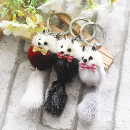 Three small plush animal keychains with long faux-fur tails and metal key rings, styled with flowers and wooden sticks on a light textured surface.