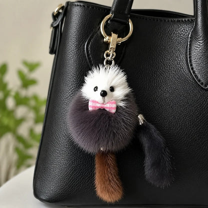 A black faux-leather handbag with a fluffy hedgehog keychain charm featuring a white and dark gray body, pink bow tie, and brown tail, hanging from a gold-tone ring.